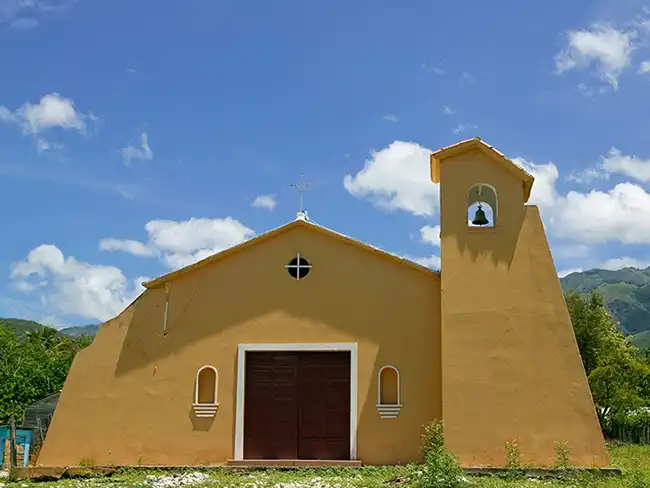 A church with a single bell in its belfry, built in the missionary style, rises from a hilltop in Baoruco Province, Dominican Republic.