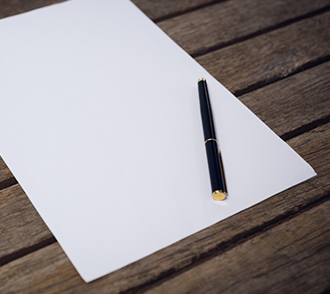 Pen and blank sheet of paper on a dark wood table.